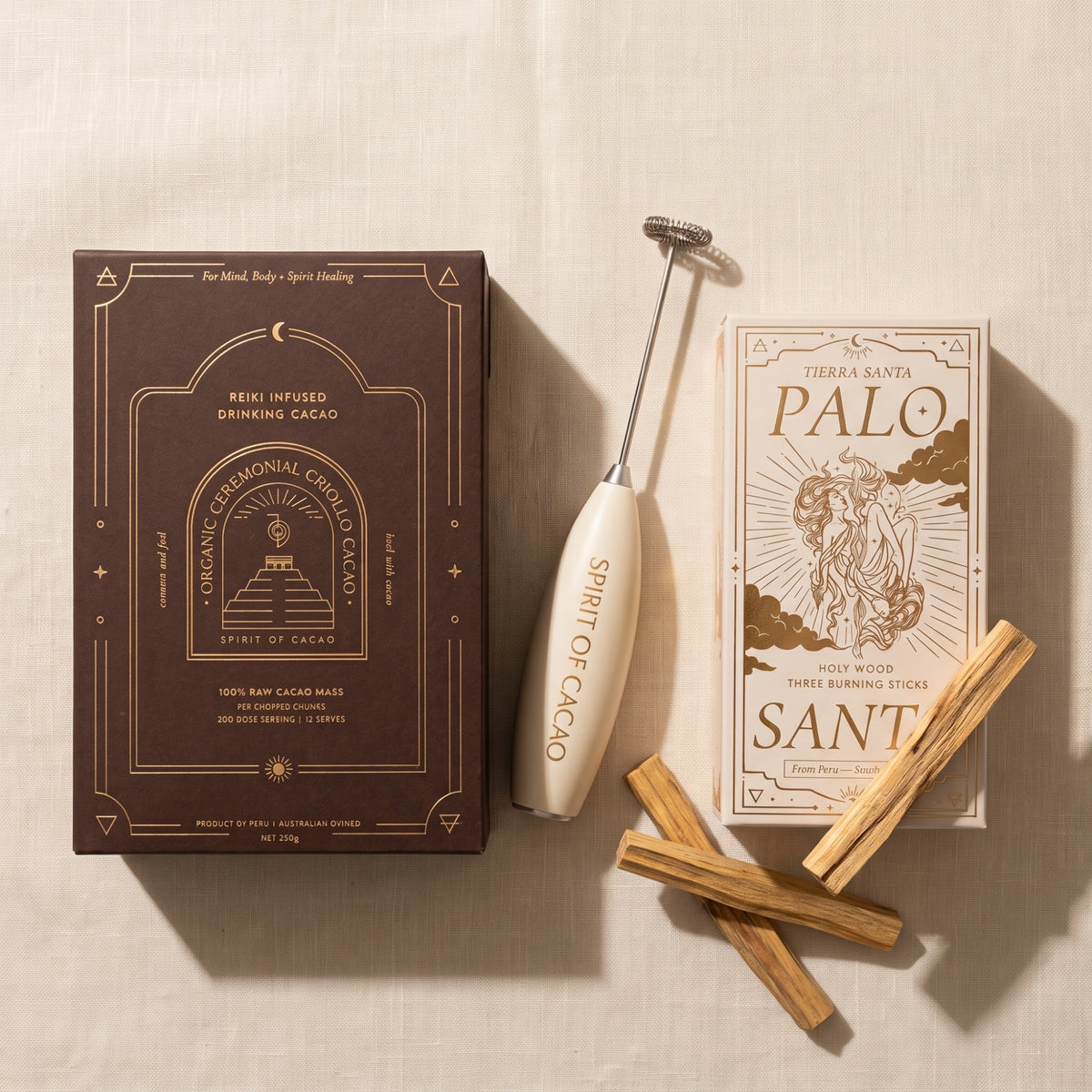 A styled flat-lay of a Reiki infused drinking cacao mix kit by Tierra Santa Palo Santo. The set includes a dark brown box of Reiki Infused Drinking Cacao, a cream-colored Spirit of Cacao handheld frother, a cream-colored Palo Santo sachet, and several natural Palo Santo wood sticks, all arranged on a textured beige fabric background.