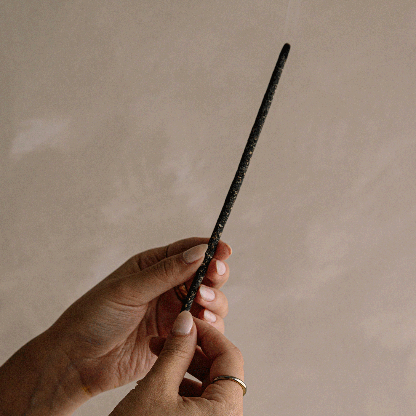 Hand holding a lit white sage incense stick with a thin trail of smoke against a soft beige wall