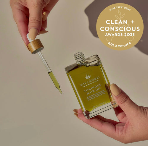 Hands holding Luminous Hair Oil bottle and dropper of golden oil beside a gold awards badge