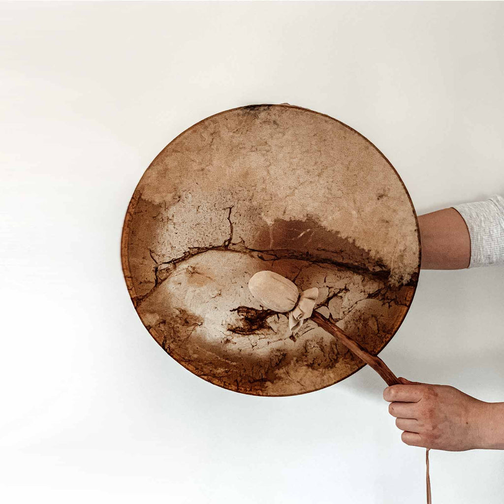 fifteen inch deer skin drum pulled over a circular pine hoop with a hand holding the drum from the back and a hand in front beating the drum with a beater made of a real tree branch and handmade topper to drum